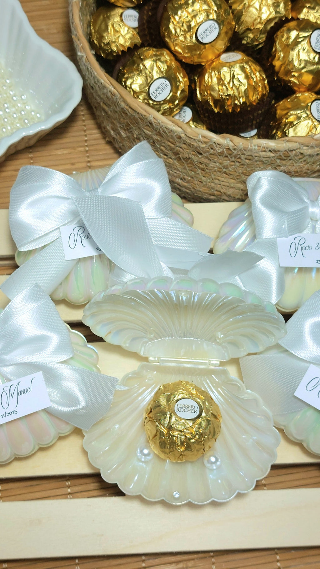 Detalle de boda con concha perlada, lazo blanco y bombón dorado personalizado con etiqueta de nombres y fecha, regalo elegante para invitados