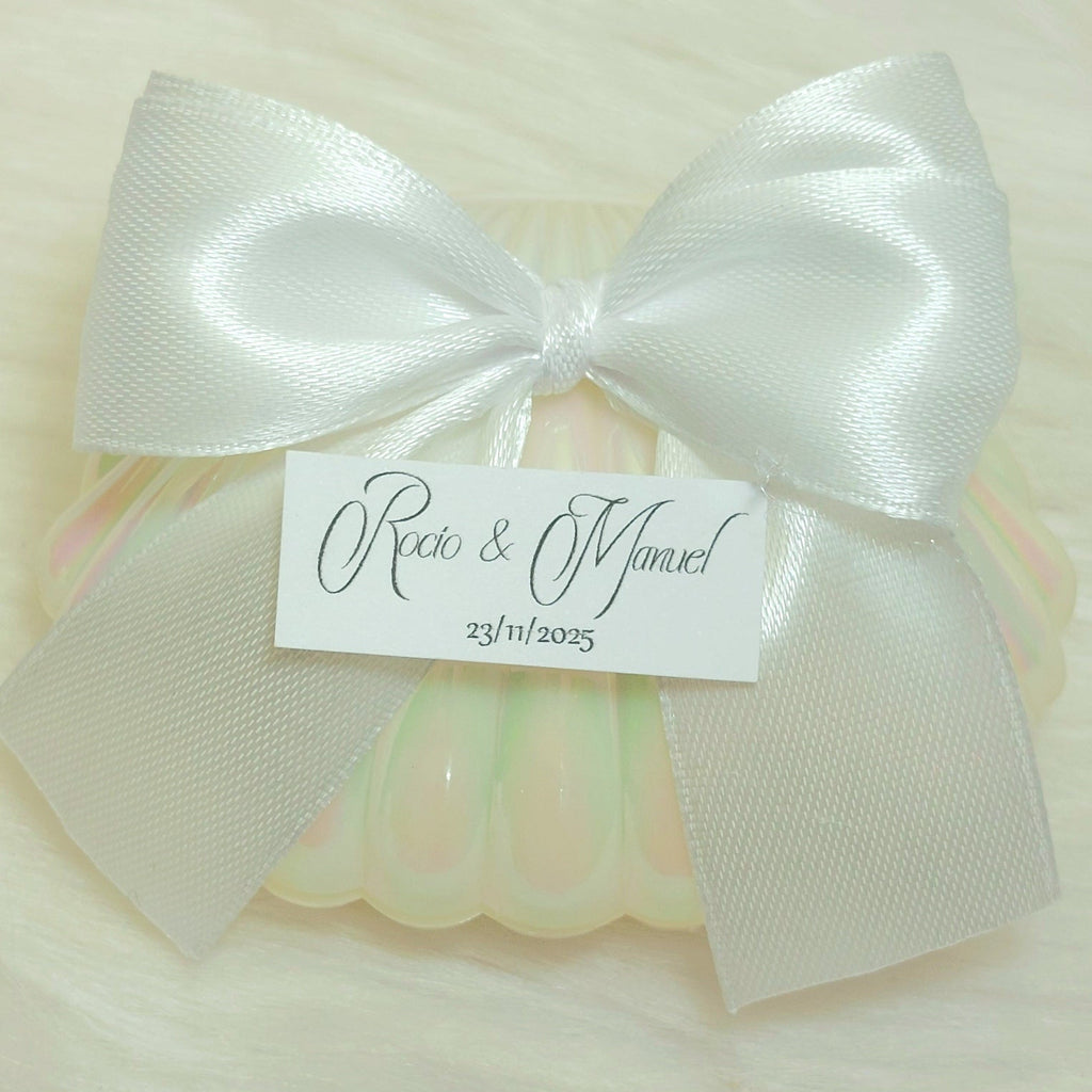 Detalle de boda con concha perlada, lazo blanco y bombón dorado personalizado con etiqueta de nombres y fecha, regalo elegante para invitados