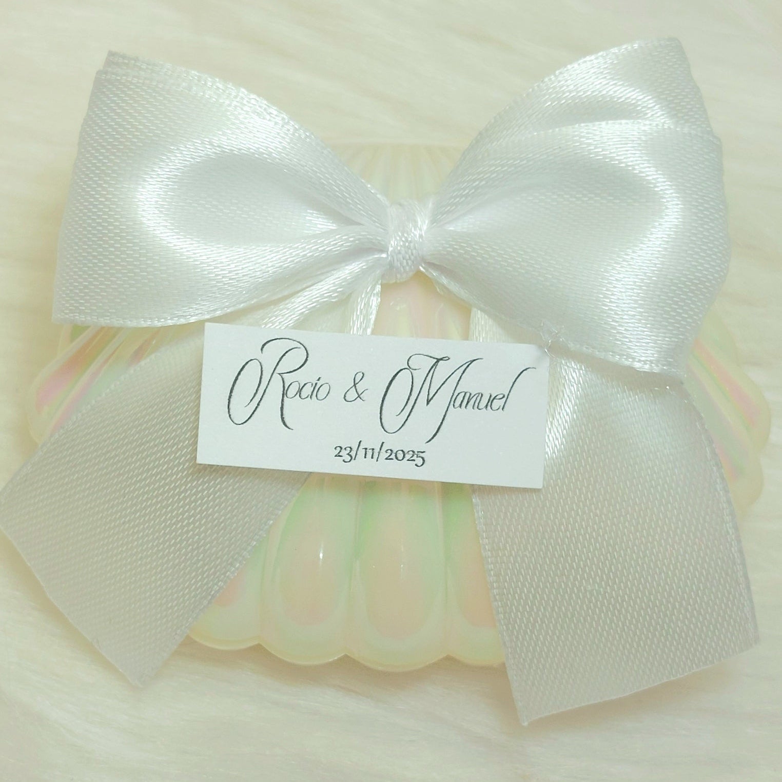 Detalle de boda con concha perlada, lazo blanco y bombón dorado personalizado con etiqueta de nombres y fecha, regalo elegante para invitados