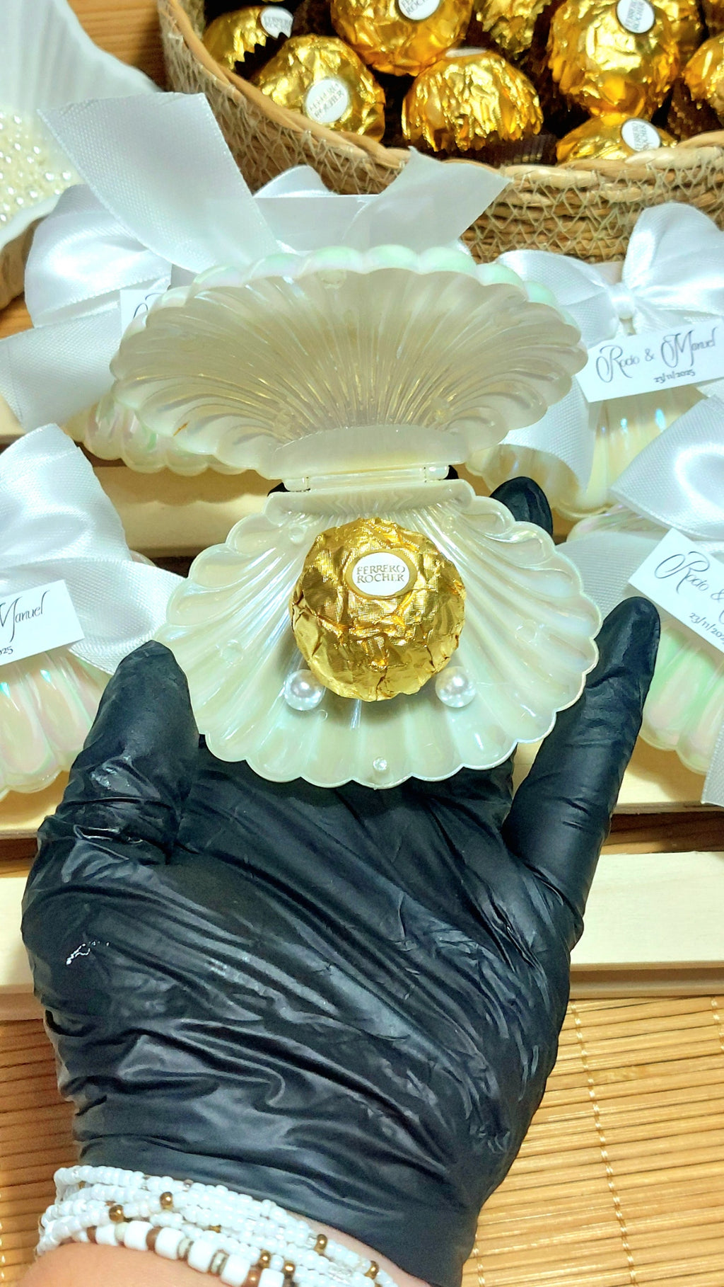 Detalle de boda con concha perlada, lazo blanco y bombón dorado personalizado con etiqueta de nombres y fecha, regalo elegante para invitados