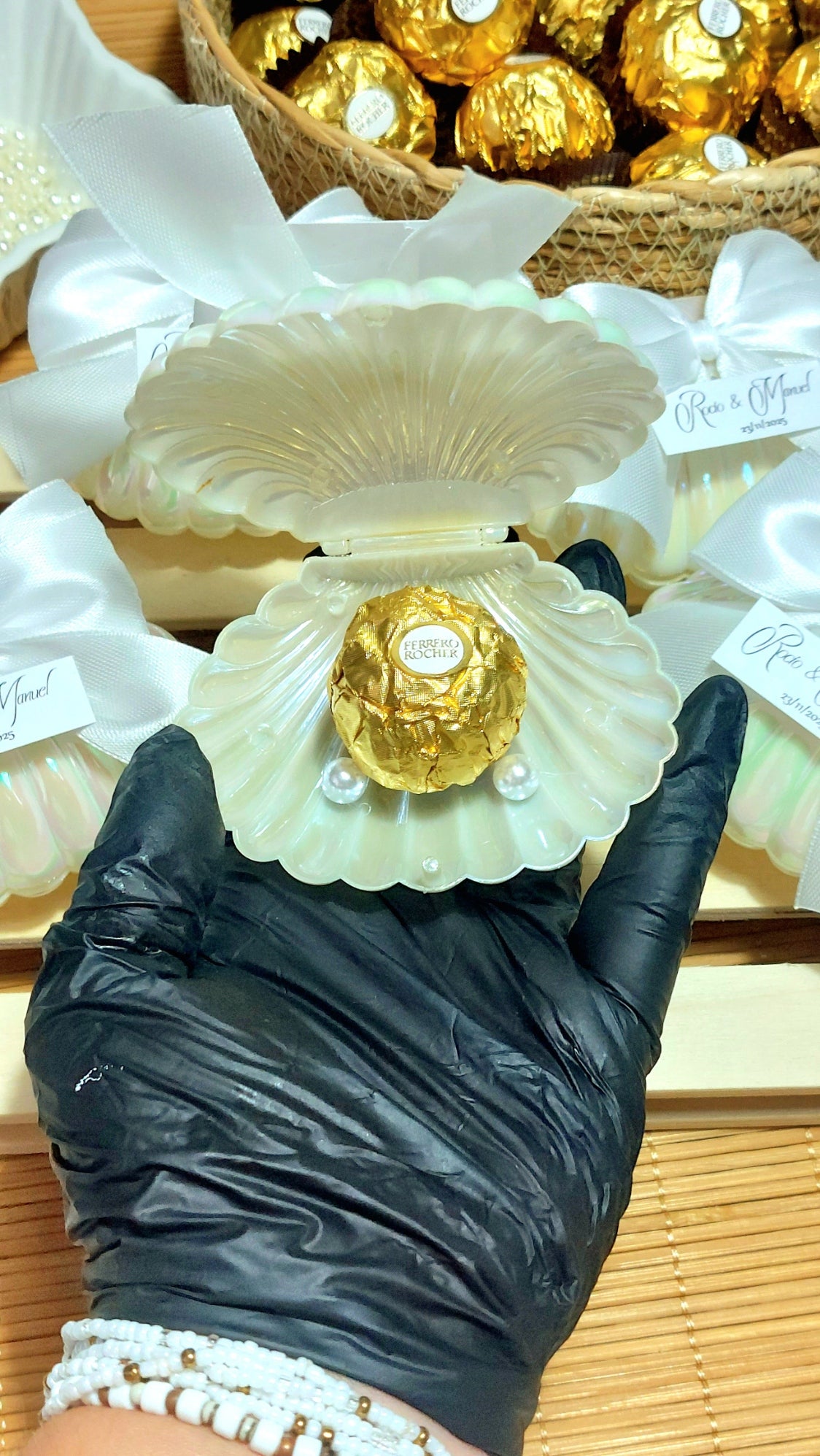 Detalle de boda con concha perlada, lazo blanco y bombón dorado personalizado con etiqueta de nombres y fecha, regalo elegante para invitados